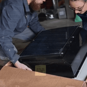 Two people lifting a solar-powered attic fan out of a box for installation into a home to cool an attic.