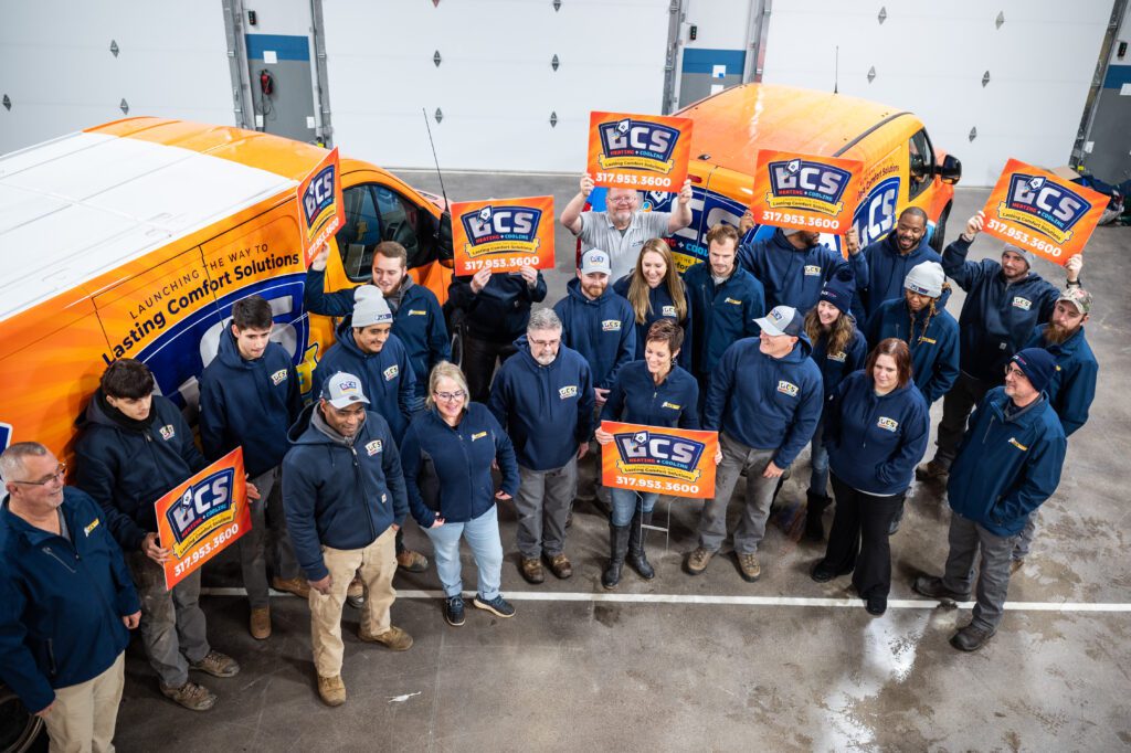 The LCS Heating and Cooling team of technicians, service professionals, office staff, and owners stand in front of LCS HVAC vehicles holding signs with the new logo in Indianapolis, Indiana